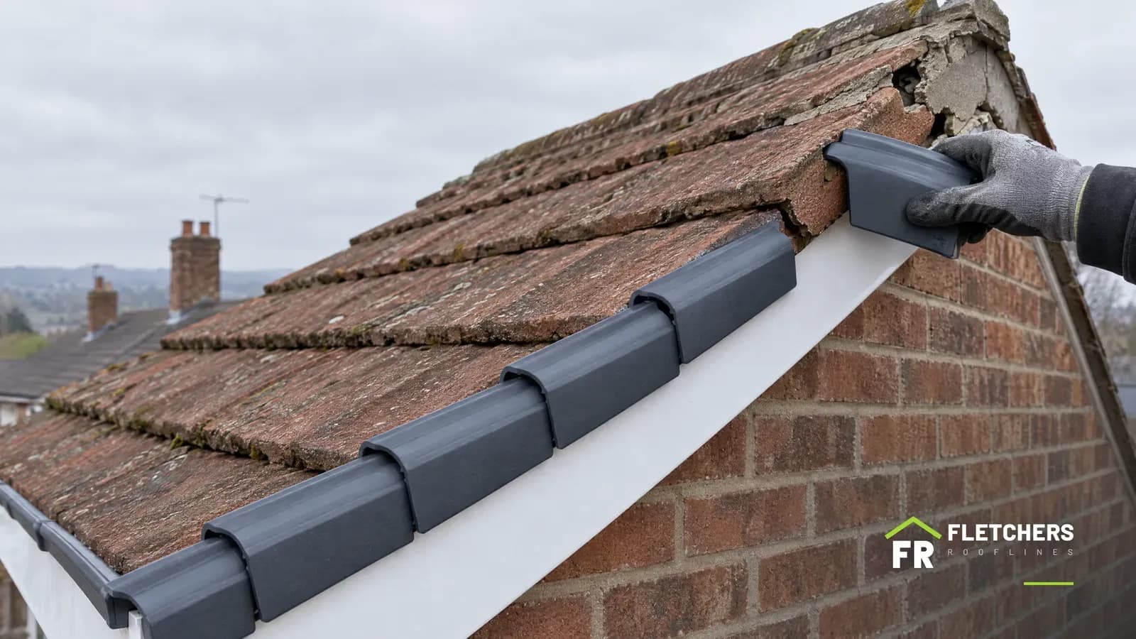 Gloved hand fitting an anthracite dry verge cap on a red tiled gable, with new caps over a white bargeboard below and old cracked mortar still at the top