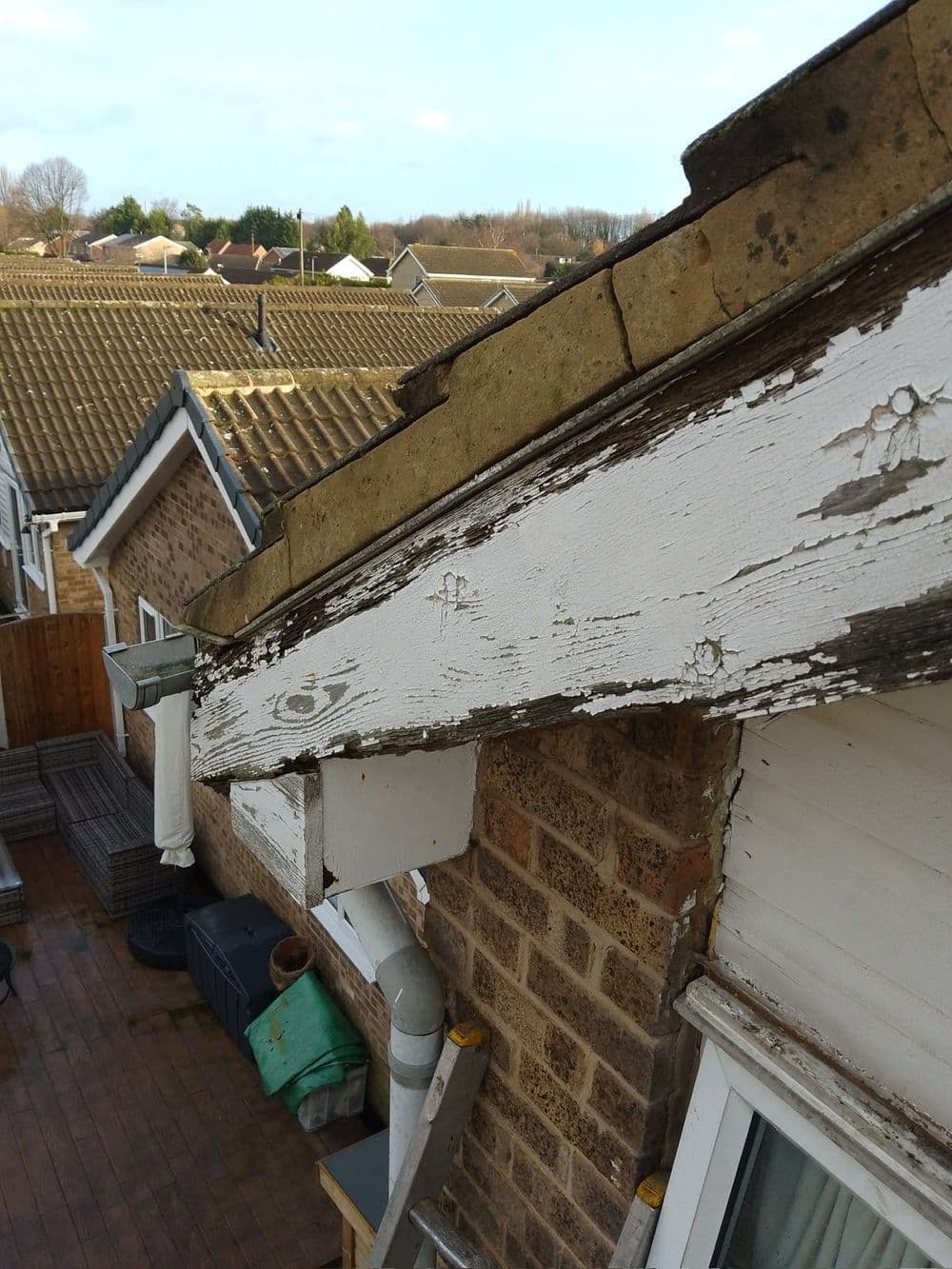 Old timber fascia with peeling white paint over a flat roof, rooftops beyond
